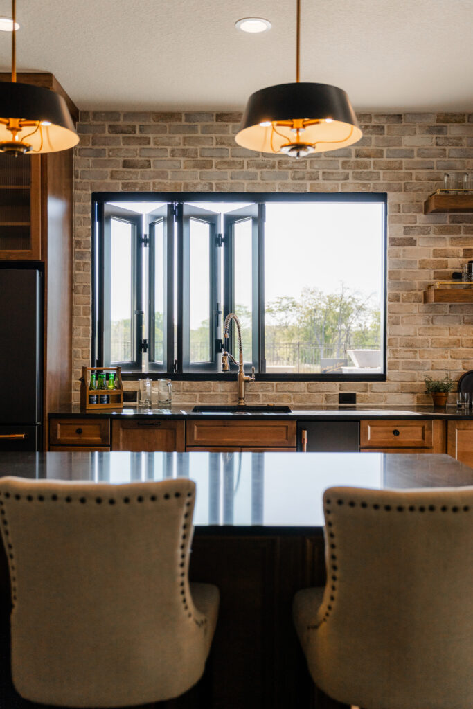 Modern kitchen with brick backsplash, open folding window, pendant lighting, and a large island with upholstered chairs.