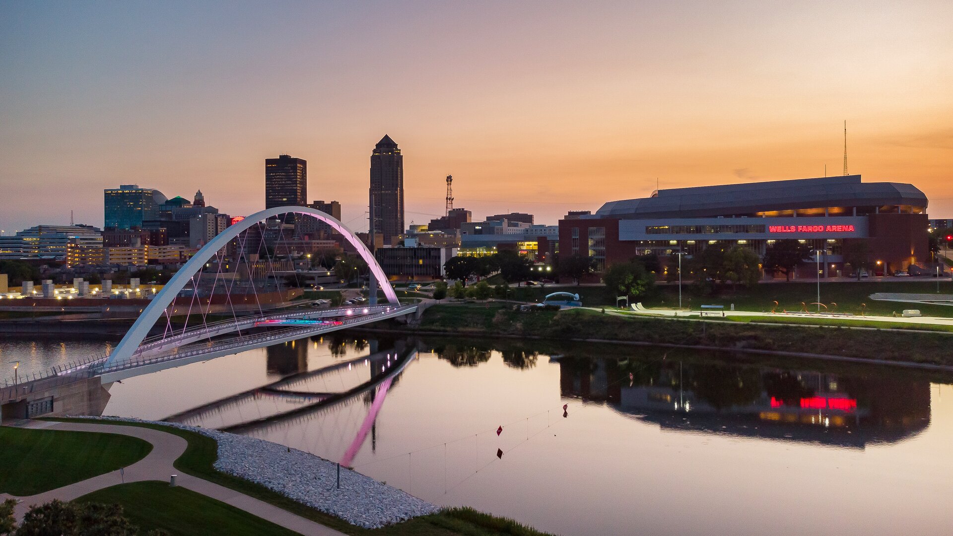 A sunset view of a modern arch bridge reflecting over a calm river, with a city skyline and an arena building in the background.