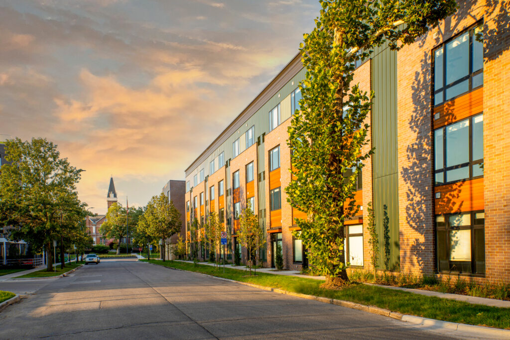 Street view of a contemporary brick and metal apartment building at sunset, lined with trees.