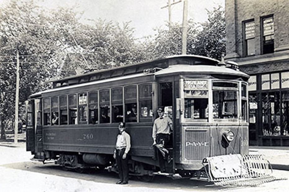 Black‑and‑white historic photo of a streetcar stopped on a city street with several people standing beside it.