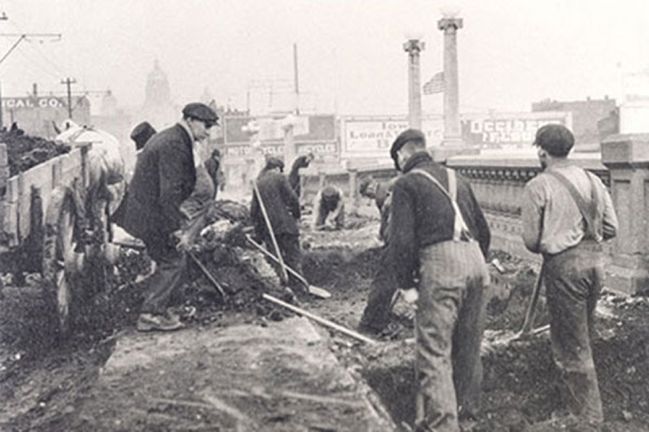Historic photo of workers using shovels to clear debris near a horse‑drawn cart on a city street.