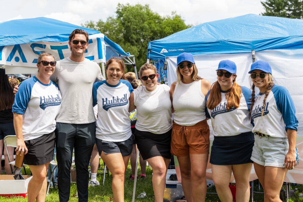A group of people standing together at an outdoor event, with tents and greenery in the background.