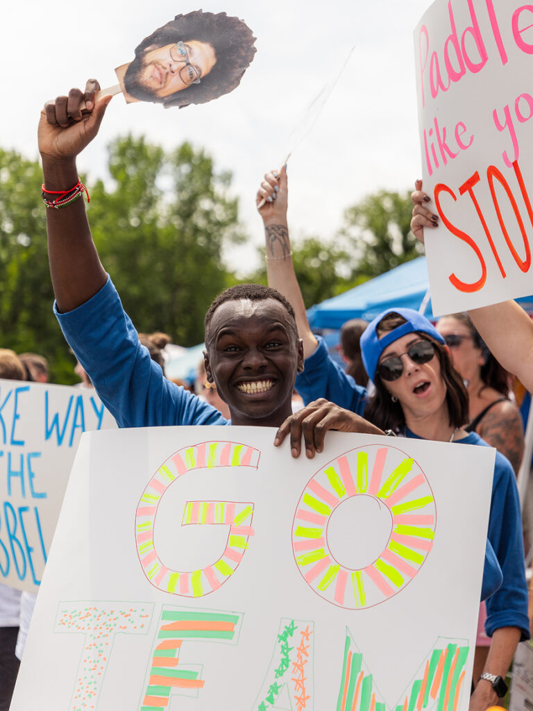 Crowd holding colorful handmade signs reading “GO TEAM” and other messages at an outdoor event.Crowd holding colorful handmade signs reading “GO TEAM” and other messages at an outdoor event.