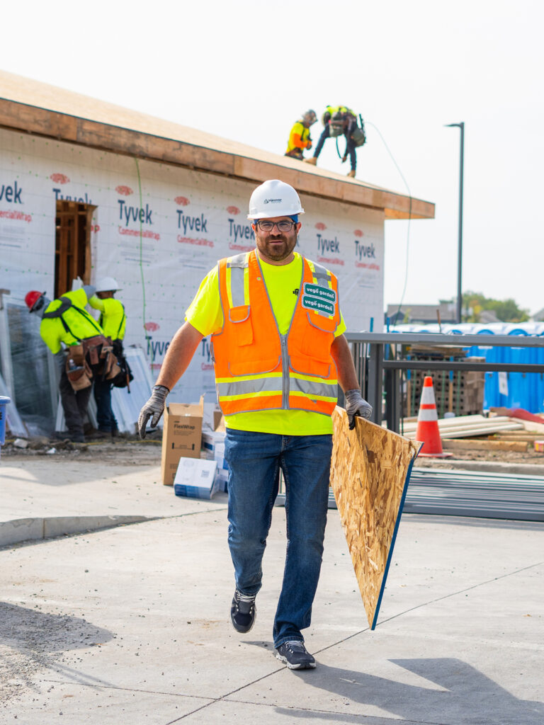 Construction site with workers, equipment, and a partially built structure covered in Tyvek wrap.