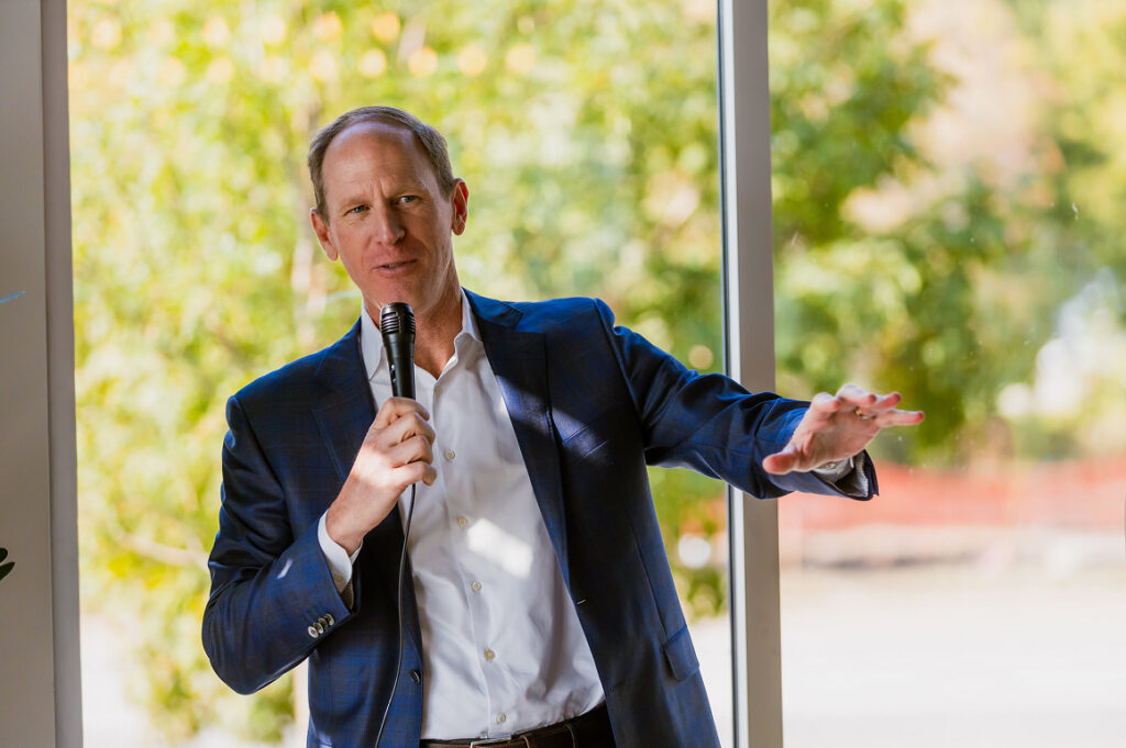 Person in business attire speaking into a microphone while standing indoors in front of large windows overlooking greenery.