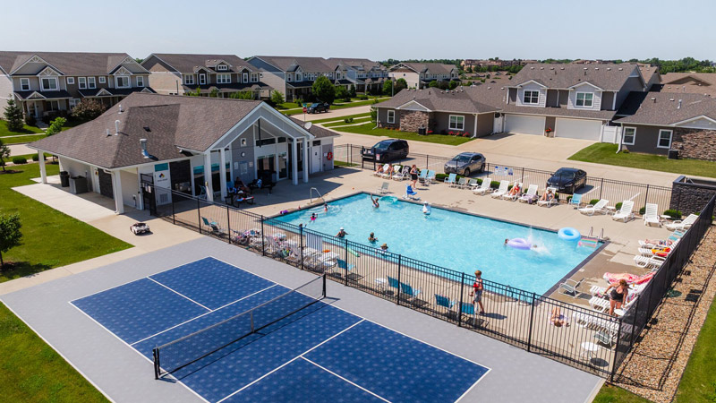 A community recreation area featuring an outdoor swimming pool with lounge chairs, a clubhouse building, nearby parked vehicles, and an adjacent sports court, surrounded by a suburban neighborhood of single‑story homes.