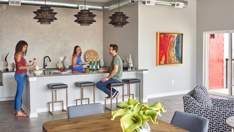 A contemporary community kitchen and lounge space with a long island, barstools, pendant lighting, modern decor, and a dining table in the foreground, with several people interacting around the counter.
