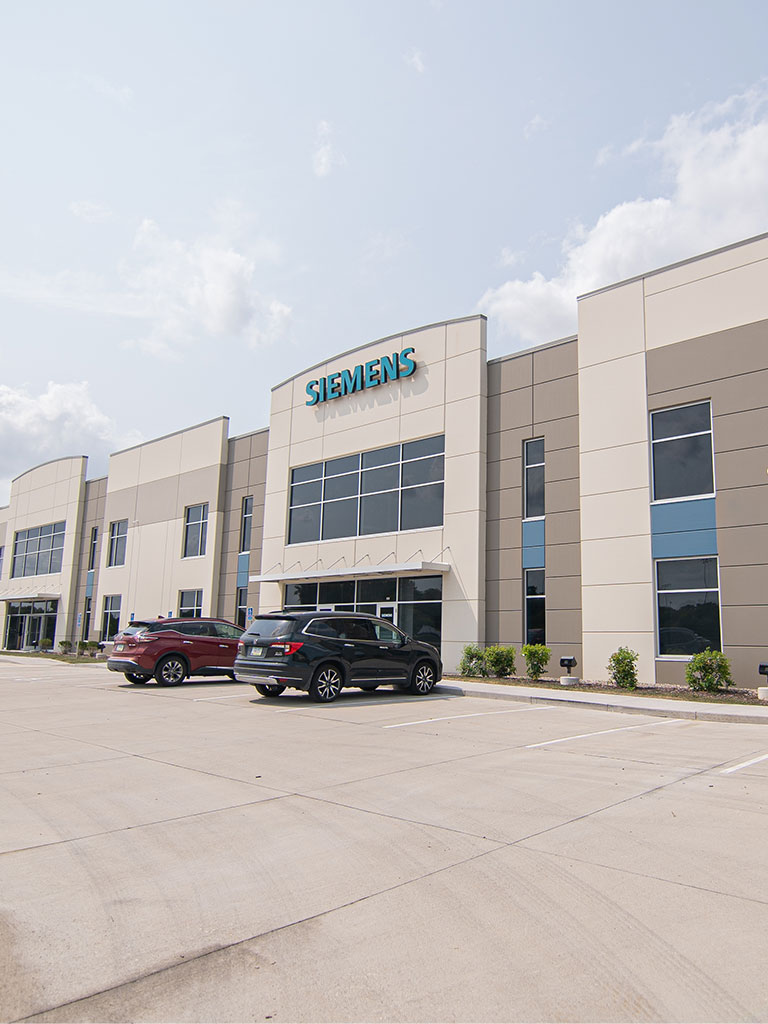 Front exterior of a modern two‑story commercial building with beige and gray facade, large windows, and cars parked in the lot; signage reading “SIEMENS” above the main entry.