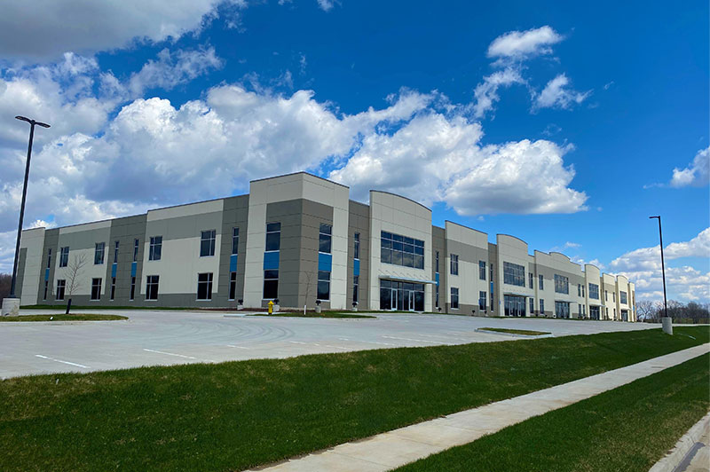 Exterior view of a large modern two‑story commercial building with beige and gray panels, large windows, and an empty parking lot under a blue sky with clouds.