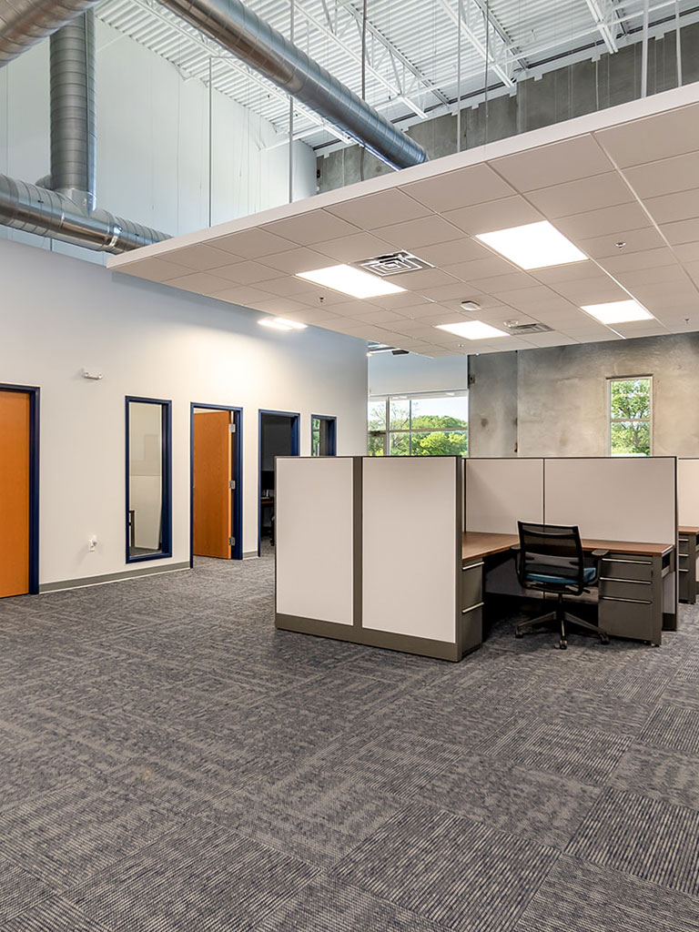 Open office area with cubicles, carpet flooring, high ceilings with exposed ductwork, and individual office doors along one wall.