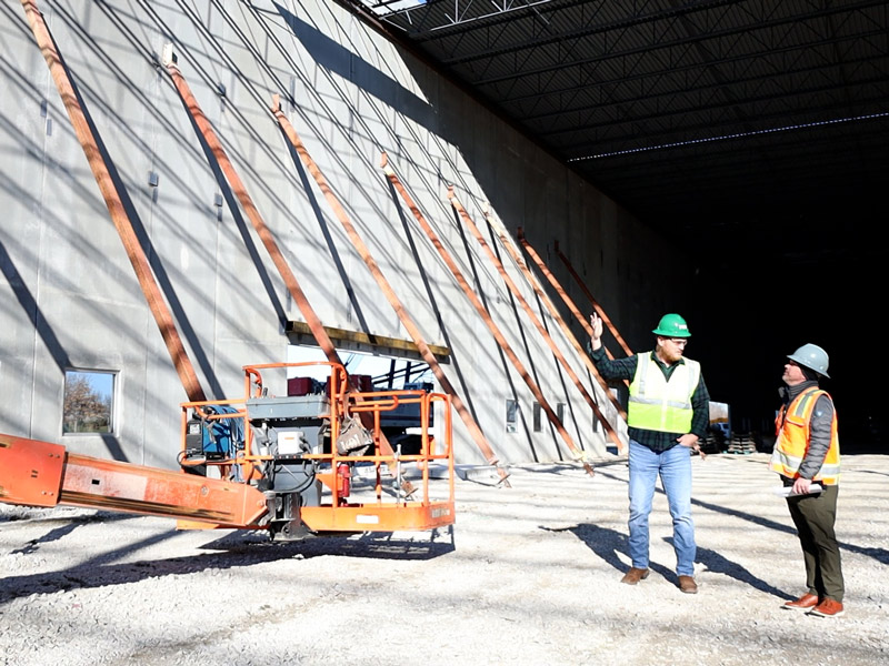Two individuals wearing hard hats and safety vests stand inside a large industrial building under construction. One person is pointing toward a tall concrete wall supported by angled braces, with construction equipment and gravel covering the ground beneath an open steel roof structure.