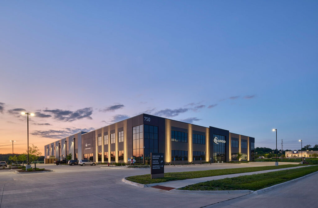 Evening exterior of a modern commercial building with illuminated windows, large parking lot, and landscaped surroundings.