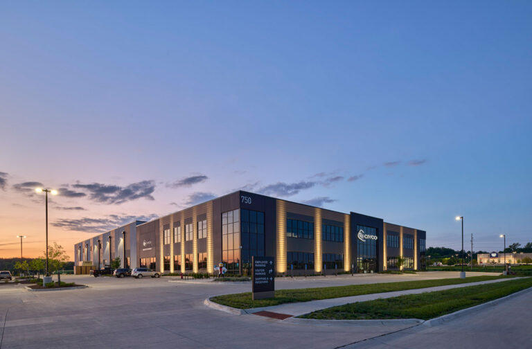 Evening exterior of a modern commercial building with illuminated windows, large parking lot, and landscaped surroundings.