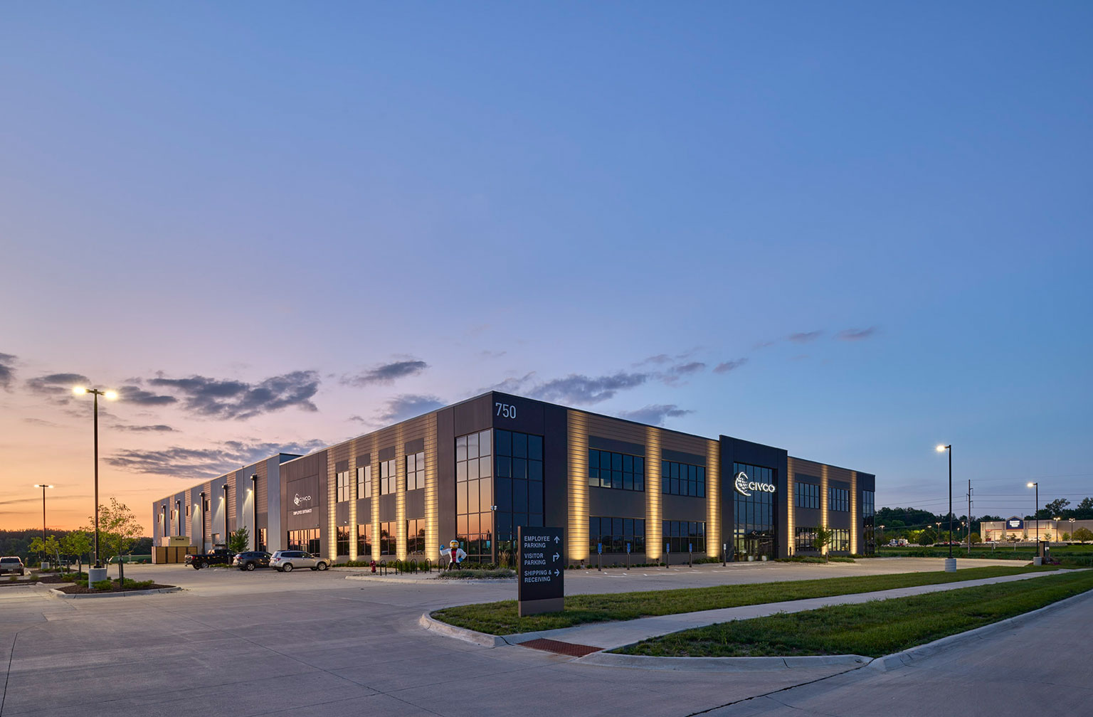 Evening exterior of a modern commercial building with illuminated windows, large parking lot, and landscaped surroundings.