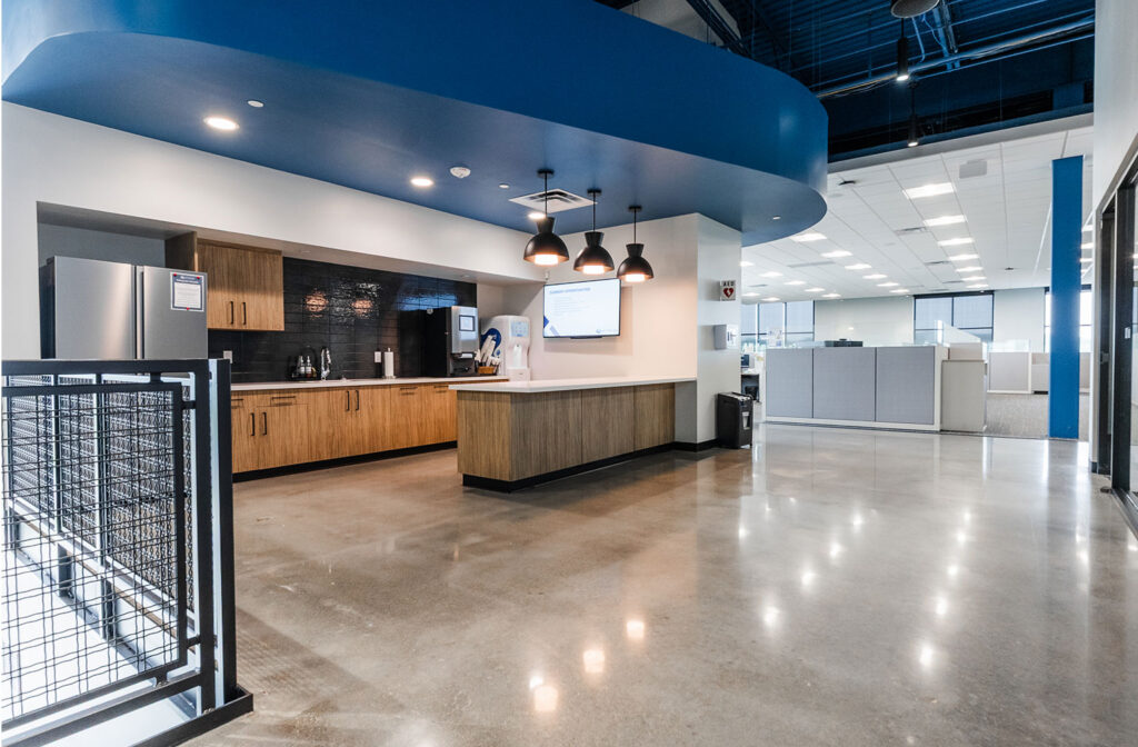 Modern office break area with wood cabinetry, stainless steel appliances, pendant lighting, and polished concrete floors next to open workstations.