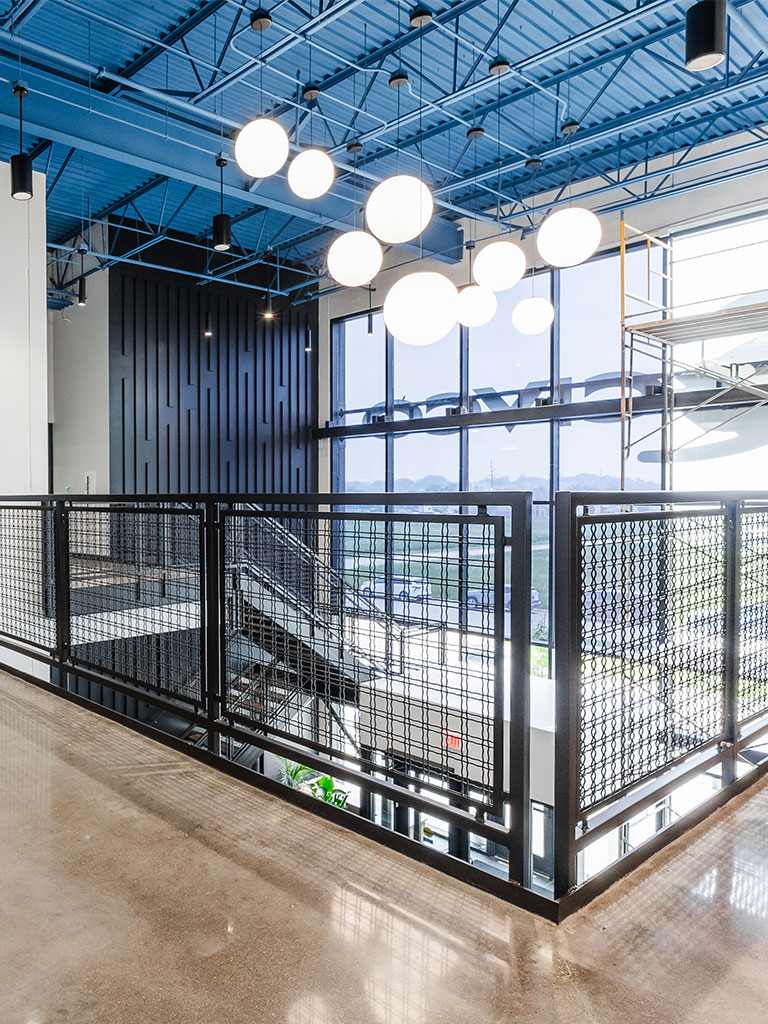 Interior view of an upper-level walkway with metal railing, large windows, blue exposed ceiling, and round hanging light fixtures.