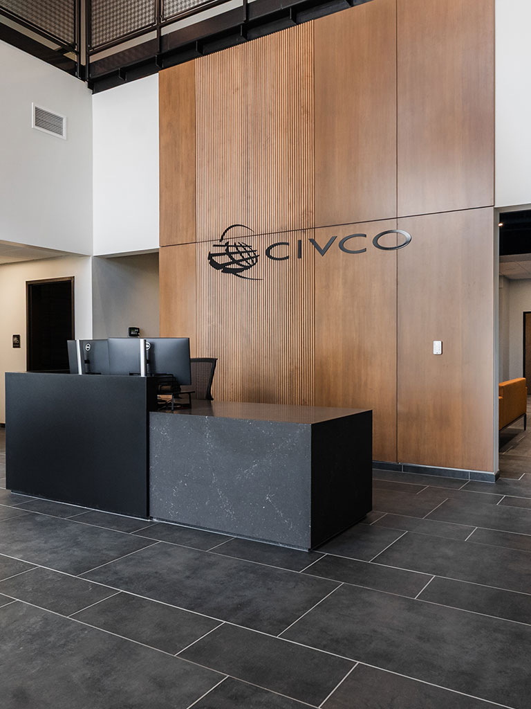 Close-up view of the CIVCO lobby reception area with a black desk, tall wood accent wall, and dark tile floors.