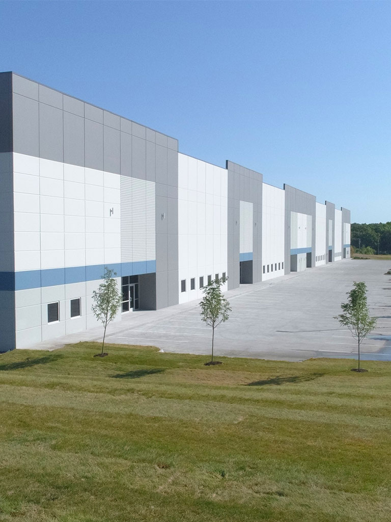 Taller view of a modern warehouse with gray and white exterior walls and a row of young trees along the concrete lot.