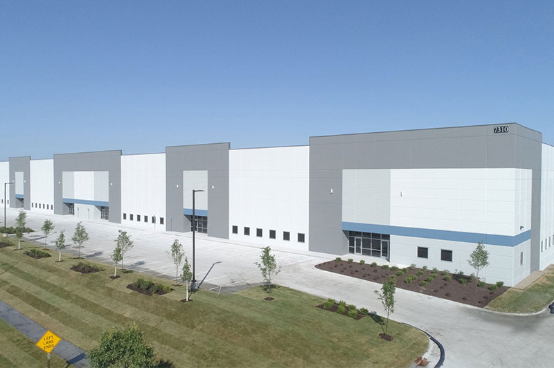 Row of white and gray warehouse bays with a sloped grassy area in the foreground and young trees lining the site.