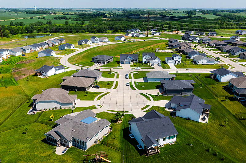 An aerial view of a suburban neighborhood featuring multiple single‑family homes arranged around a circular street layout, surrounded by large green lawns, open fields, and distant trees under a clear sky.