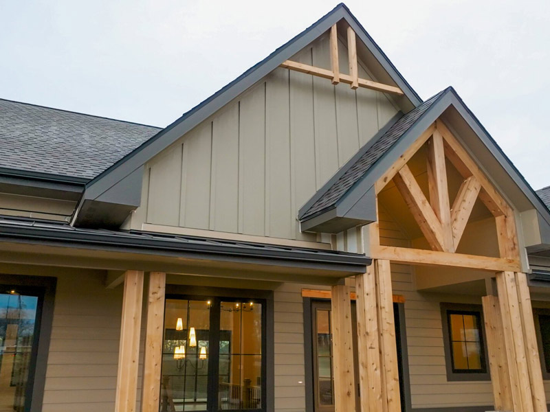 Exterior view of a craftsman‑style home featuring board‑and‑batten siding, timber posts, and large windows with warm interior lighting visible.