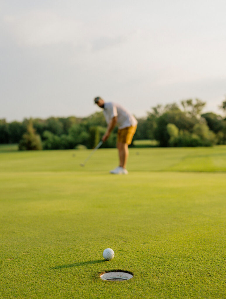 A golf ball rests near the edge of a hole on a green, with a golfer in the background preparing to putt on a sunny course.