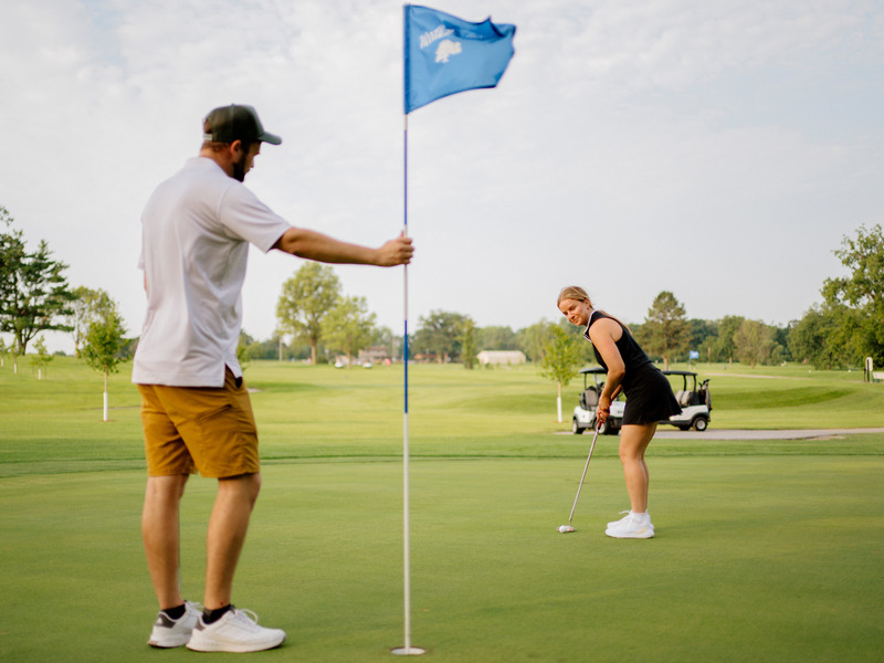 Two people stand on a golf green, with one holding the flagstick beside the hole while the other prepares to putt. A golf cart and trees are visible in the background under a partly cloudy sky.