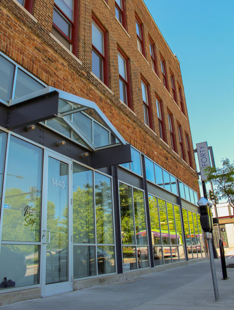 Street-level view of a brick building entrance with large glass windows and metal awnings.