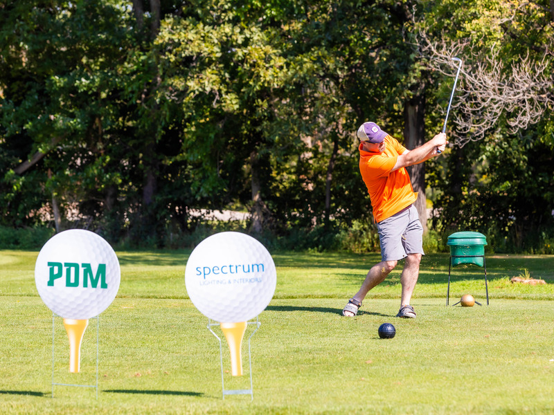 A person swings a golf club on a sunny course, with two large golf‑ball‑shaped sponsor signs labeled “PDM” and “spectrum” displayed in the foreground.