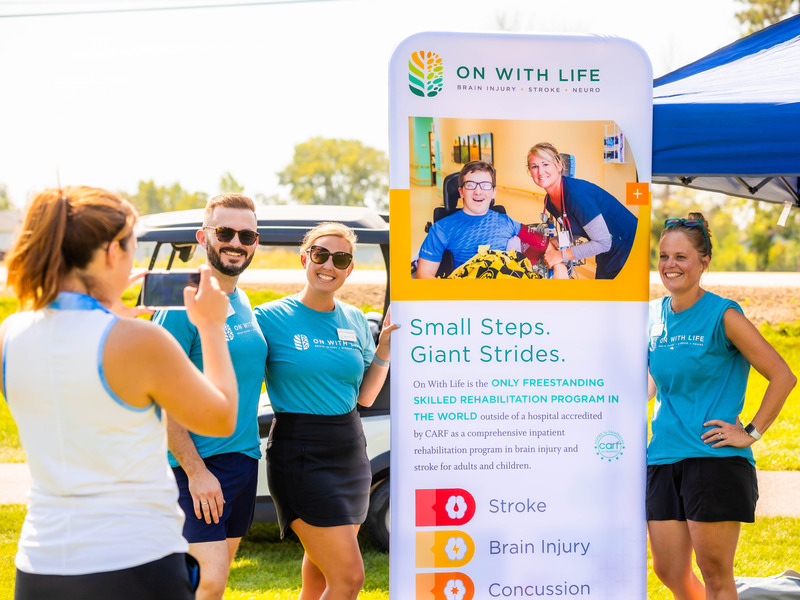 Hubbell team members wearing matching teal shirts stand near a vertical event banner for “On With Life,” which highlights brain injury, stroke, and neuro rehabilitation services at their annual extreme charity golf classic.