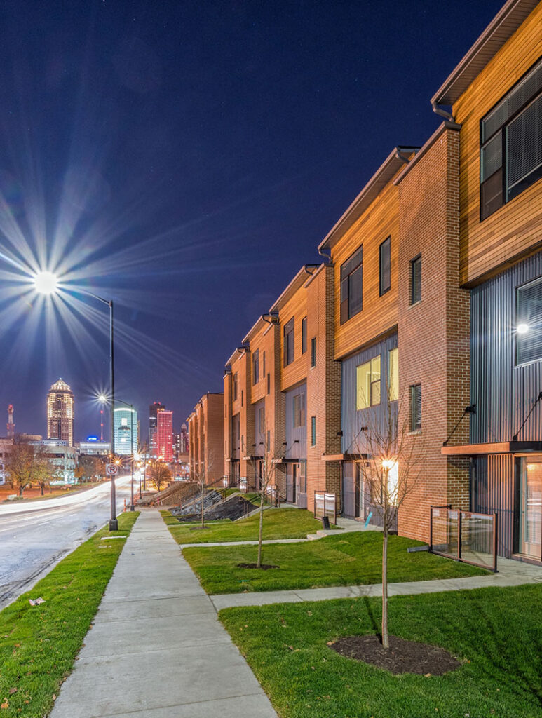 Modern townhomes lining a sidewalk at night with city skyline lights in the distance.