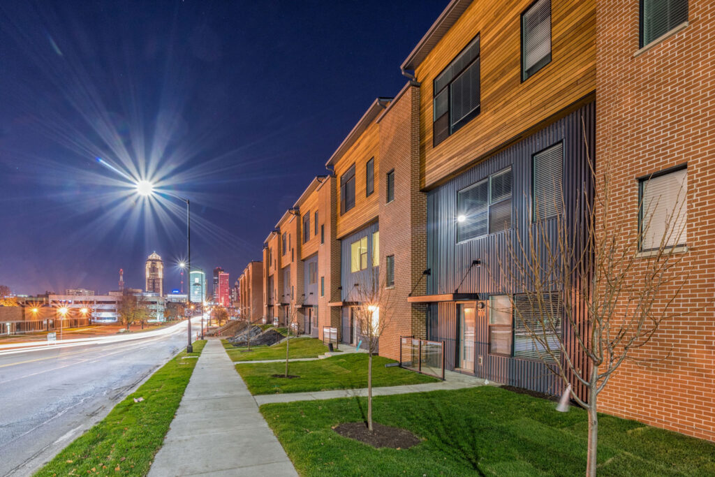 Night view of modern townhomes along a landscaped sidewalk with nearby city skyline.