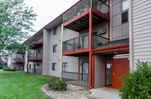 Exterior of a multi‑story Hubbell apartment building with balconies, gray siding, red trim, and a landscaped lawn and walkway in front.