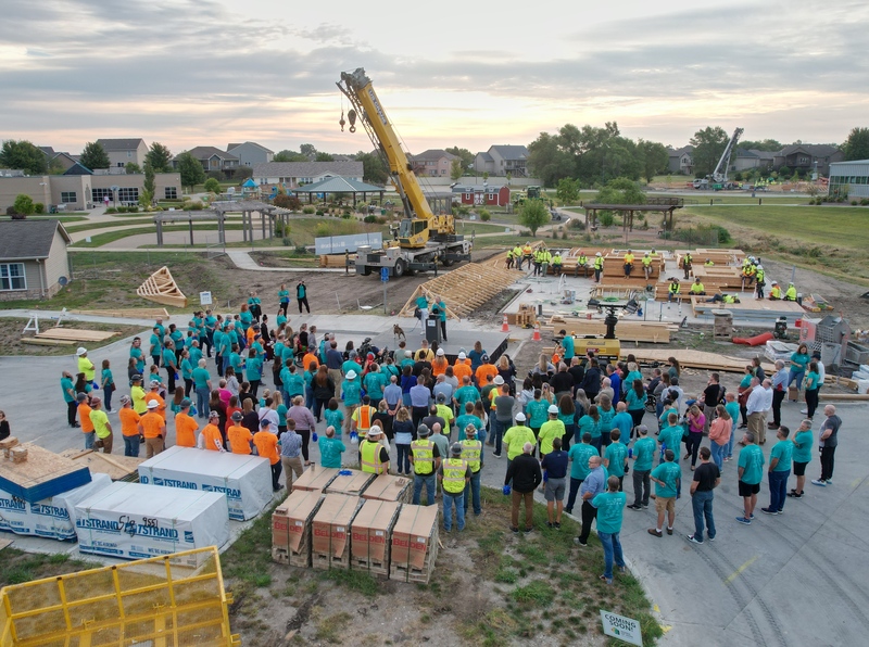 A large group of volunteers gathers at a neighborhood construction site with a crane and building materials nearby.