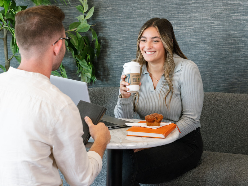 Two people sit at a small round table in a café-style setting, one holding a coffee cup and the other holding a notebook. A laptop, notebook, and a muffin are on the table, with greenery visible in the background.