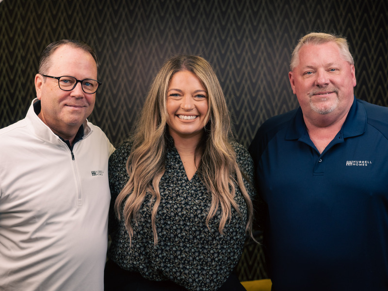 Three people stand closely together in front of a patterned dark wall, with two wearing collared shirts and one wearing a patterned top with long wavy hair.