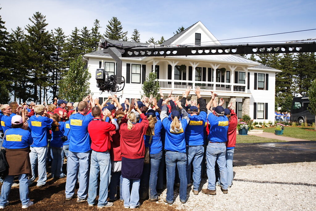 A crowd in matching shirts raises their hands toward a camera crane in front of a large white house with a wraparound porch during ABC's Extreme Makeover.