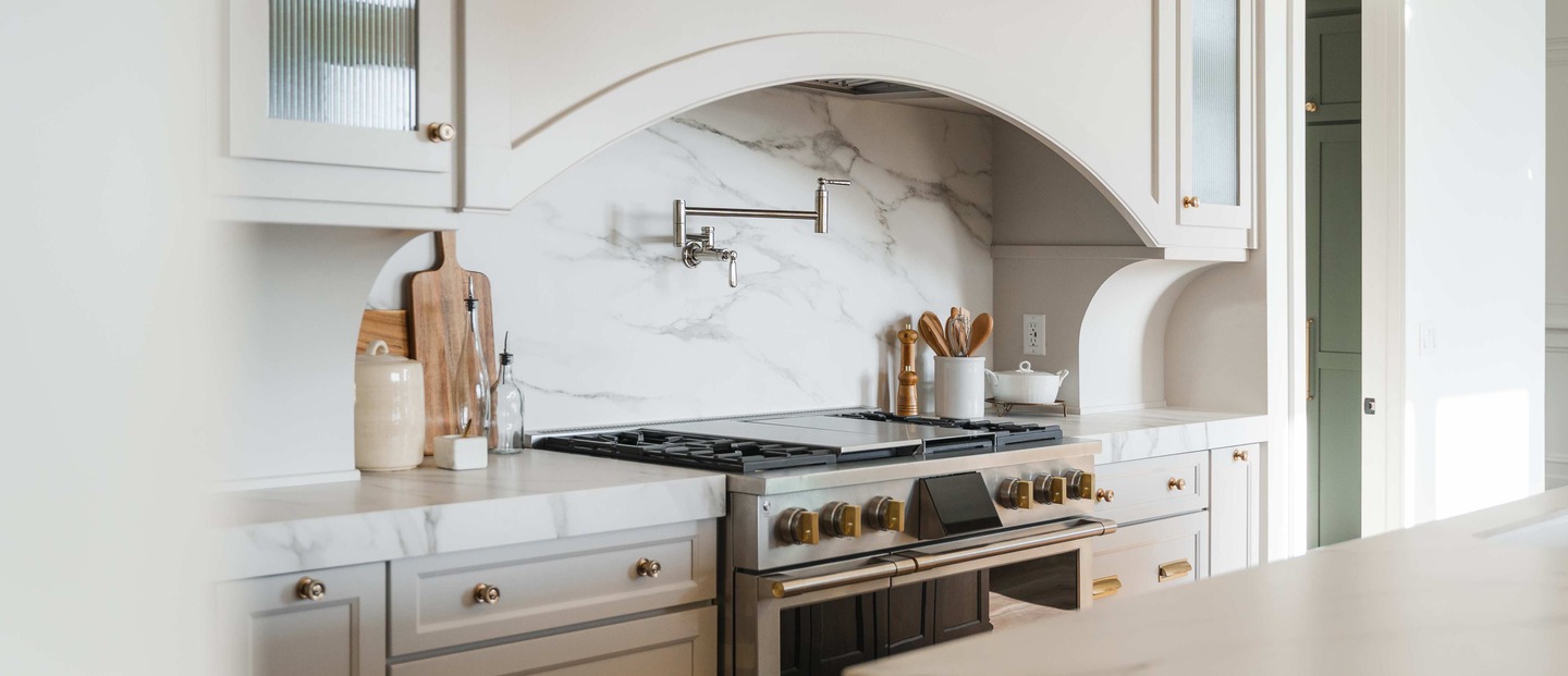 Modern kitchen with a stainless steel stove, gold accents, marble backsplash, and light cabinetry with simple countertop decor.