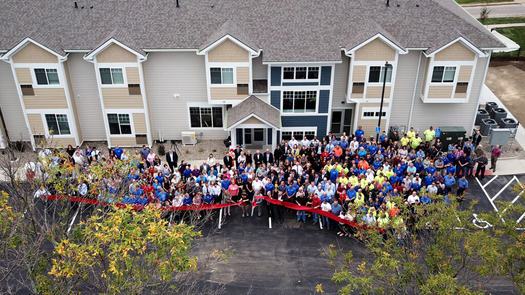 An overhead view of a large group of people gathered in front of the newly constructed Ronald McDonald House of Central Iowa, standing behind a long red ribbon for a ribbon‑cutting event.