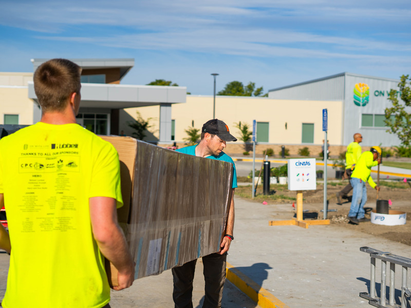 Workers carry a large boxed item across a construction site near a modern building, while other crew members work in the background.