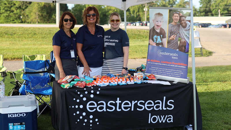 Three people stand behind a table under a canopy at an outdoor event, displaying snacks and an Easterseals Iowa information board.