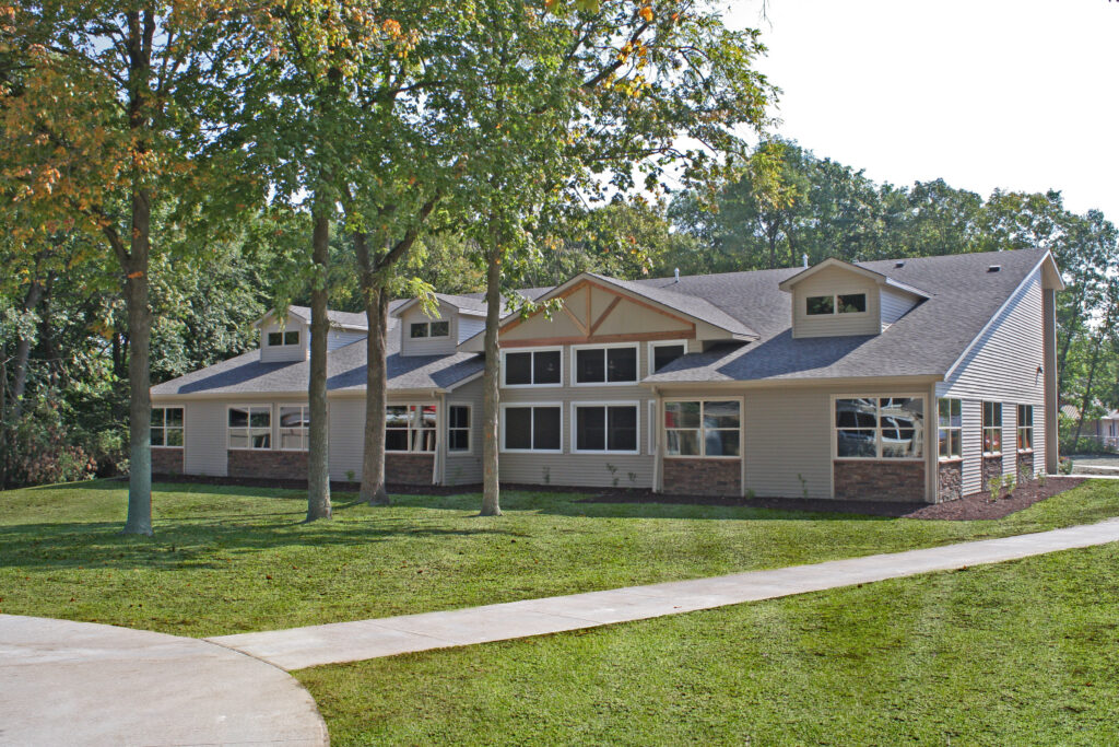Hubbell's Easter Seals Camp Sunnyside in the “Cabins for Campers” single‑story building with large windows, tan siding, and brick accents sits behind several tall trees, surrounded by green lawn and a curved sidewalk.