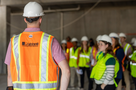 A person wearing a hard hat and an orange safety vest with the Hubbell Construction Services logo stands facing a group of people in safety gear during a tour or briefing inside an unfinished construction area.