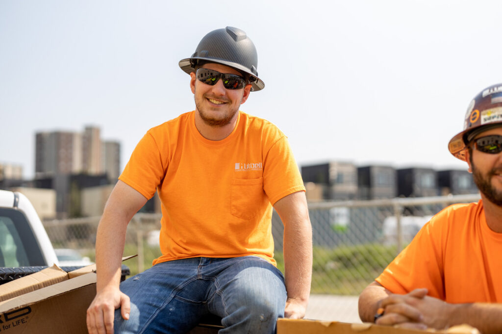 A person in an orange construction shirt and hard hat sits on the back of a vehicle or work surface at a construction site, with buildings and fencing visible in the background. Another worker in similar safety gear is partially visible on the right side of the image.
