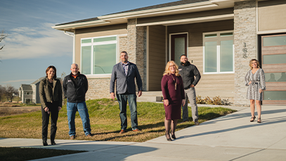 A group of six Hubbell team members stand spaced out on the driveway and lawn in front of a modern single‑story home with stone and siding exterior.