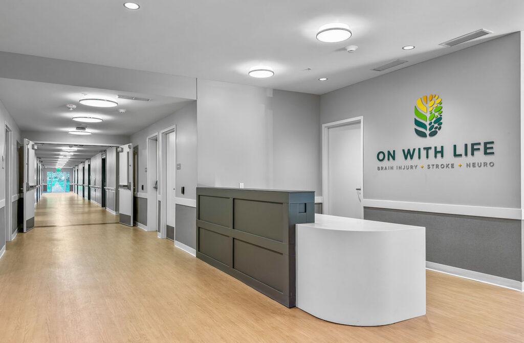 Interior hallway and reception at the Hubbell‑built On With Life Long Term Skilled Care Facility with wood flooring and the On With Life logo on the wall.