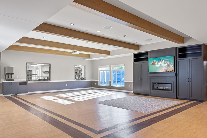 Spacious multipurpose room in the Hubbell‑built On With Life Long Term Skilled Care Facility with wood flooring, natural light, and built‑in media cabinetry.