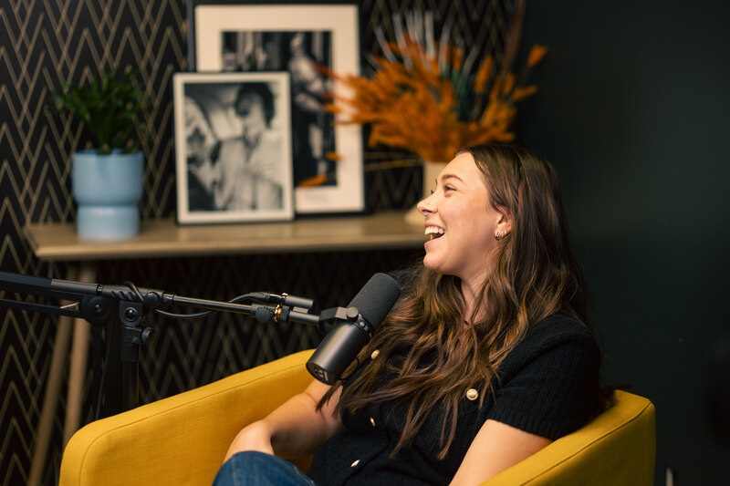 Person seated in a yellow chair speaking into a podcast microphone, with framed artwork, a plant, and decorative background elements behind them.