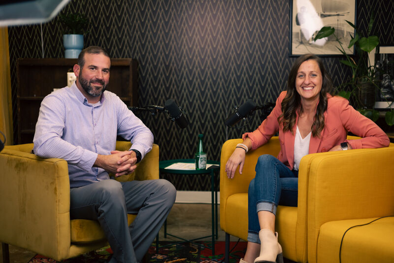 Two people seated in yellow armchairs in a podcast studio setup with microphones, a patterned dark wall, framed artwork, shelving, and plants in the background.