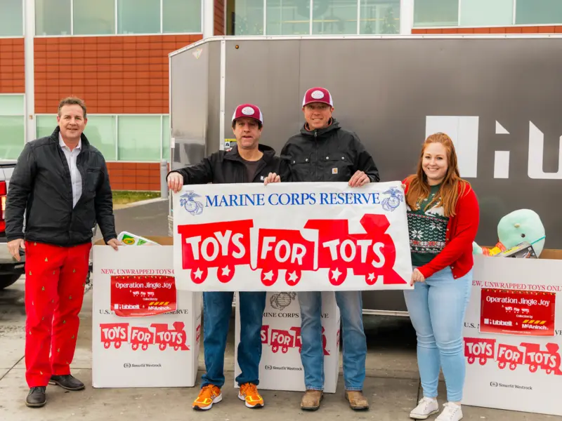 A group of people stand in front of a trailer holding a large ‘Marine Corps Reserve Toys for Tots’ banner, with donation boxes filled with toys surrounding them.